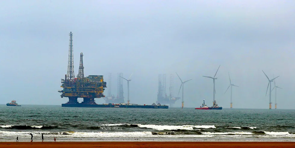 A Shell platform is towed for decommissioning surrounded by offshore wind turbines.