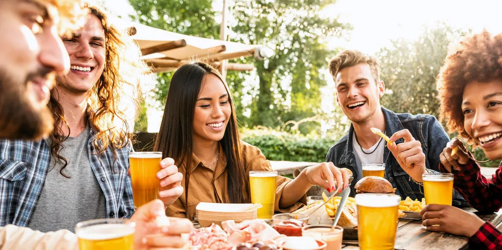 Group of multiracial friends having backyard dinner party together - Diverse young people sitting at bar table toasting beer glasses in brewery pub garden - Happy hour, lunch break and youth concept . restaurant diners foodservice.