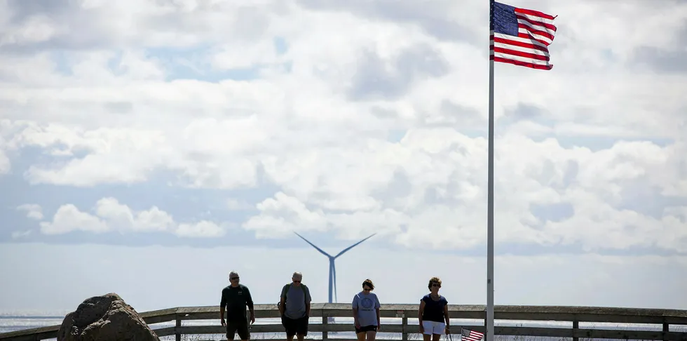 Tourists walk past an American flag on Rhode Island's Block Island, with one of the eponymous 30MW wind farm's turbines in the background