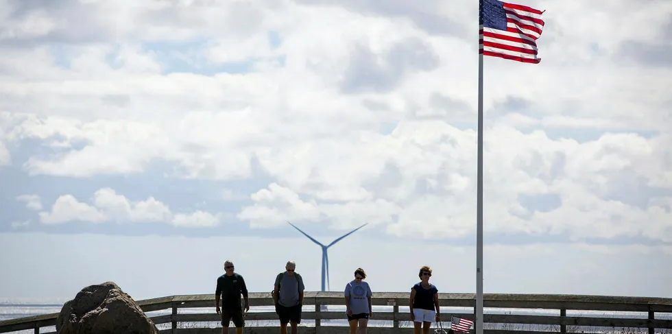 Tourists walk past an American flag as a GE wind turbine stands off Block Island, Rhode Island, US.