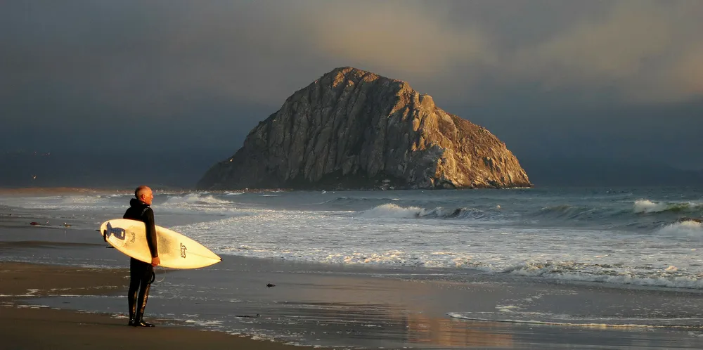 A surfer looks out at the Pacific Ocean as Morro Rock stands under evening sun on July 8, 2014 in Morro Bay, California.