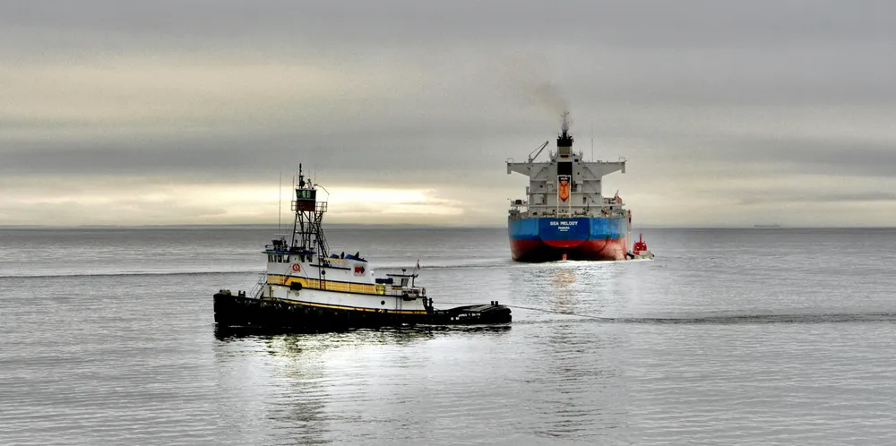 A tug is seen in the foreground as a ship steams away. Emissions are seen from the ship.