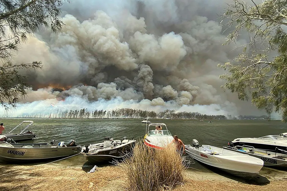Brannrøyk dekker himmelen over småbåter som er trukket på land ved Lake Conjola i Australia torsdag. Turister evakueres fra utsatte steder sørøst i landet. Foto: Robert Oerlemans / AP / NTB scanpix