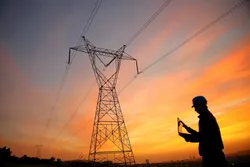 An electrical engineer with a laptop near a high-voltage transmission pylon.